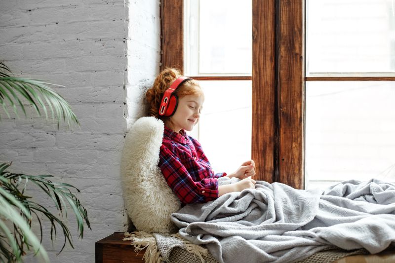 A teenage girl with red curly hair enjoys listening to music sitting on the window sill